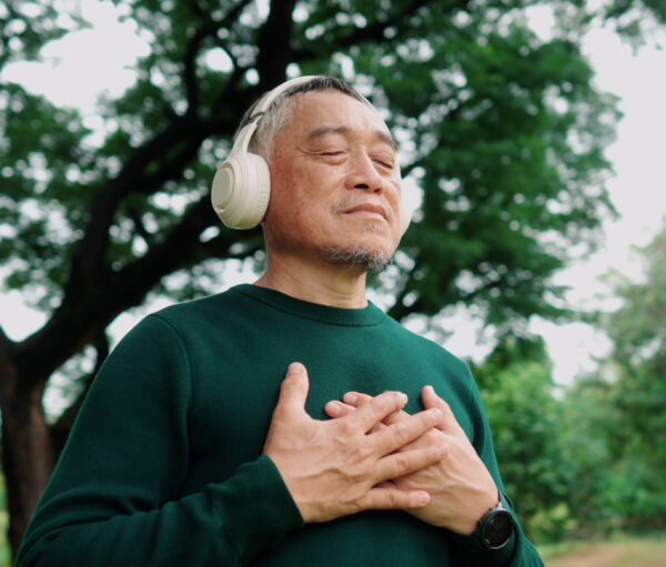 Elderly Asian man meditating under a tree with wireless headphones, calm and refreshed.