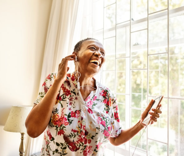 Cheerful senior female standing by window enjoying listening to music on earphones