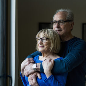 A photo of thoughtful senior couple looking through window. Loving man embracing woman at home. They are wearing casuals.